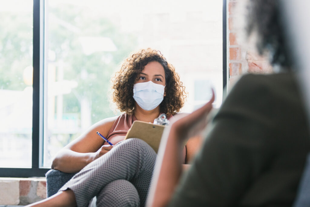 A confident female counselor attentively listens as a patient discusses something during a session. The counselor is wearing a protective mask as she is working during the COVID-19 pandemic.